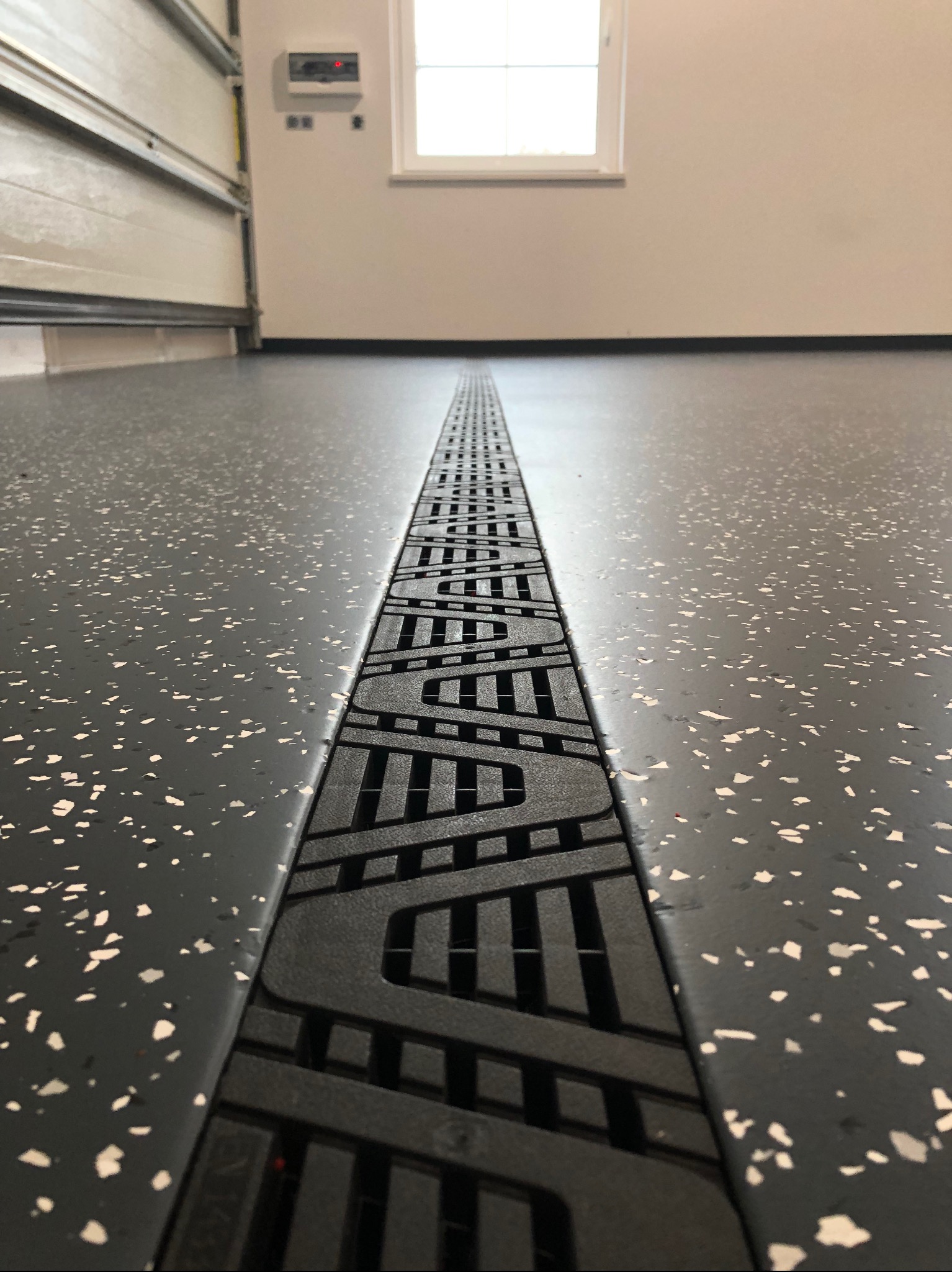 Szara posadzka żywiczna z odpływem liniowym w garażu. Widoczna brama garażowa i okno. Detal odpływu z wzorem. Nowoczesne wykończenie podłogi.