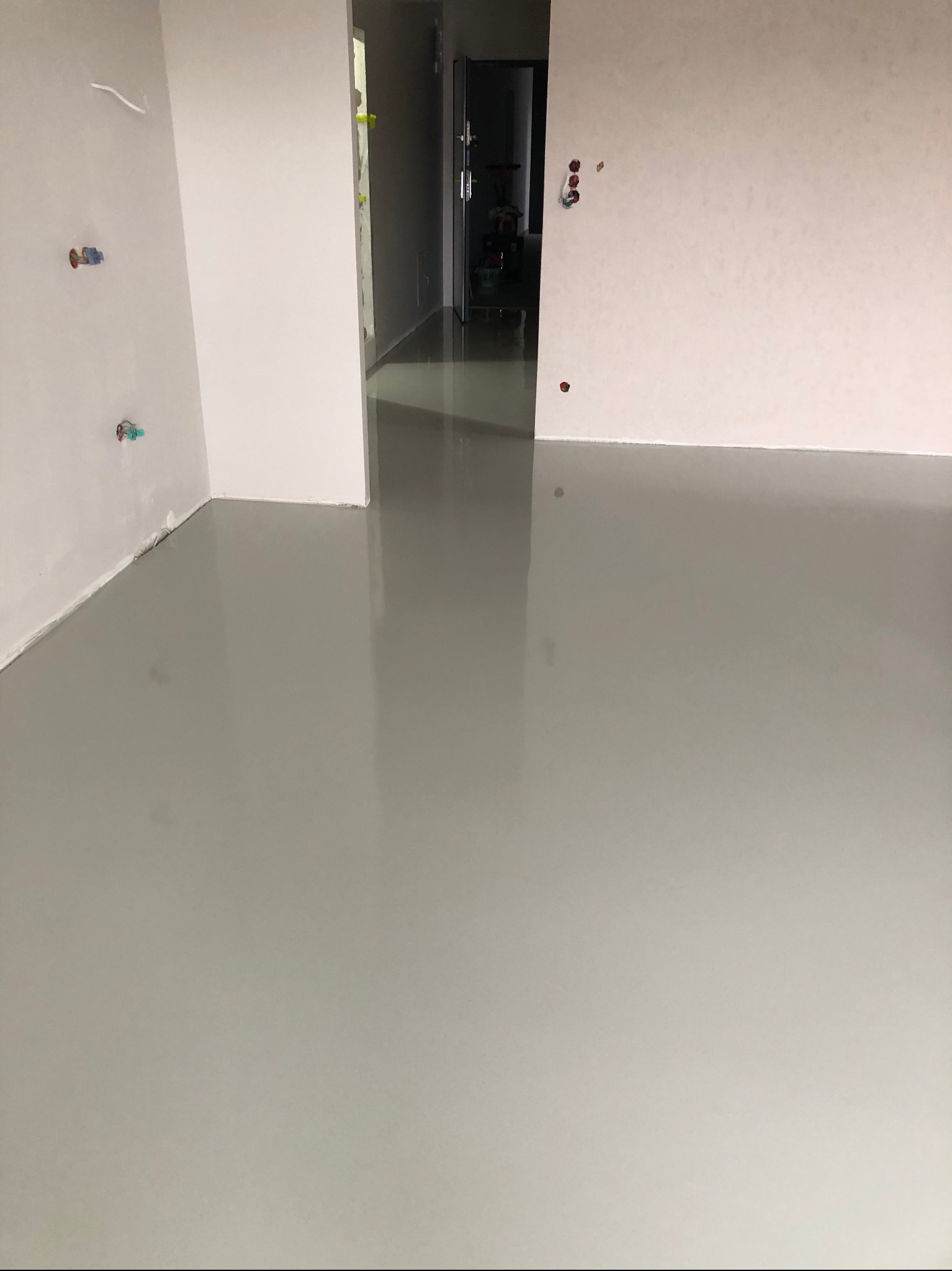 Jasnoszara, gładka posadzka w pustym pomieszczeniu z widocznymi instalacjami na ścianie i otwartym przejściem do kolejnego pokoju. Błyszcząca powierzchnia odbija światło.