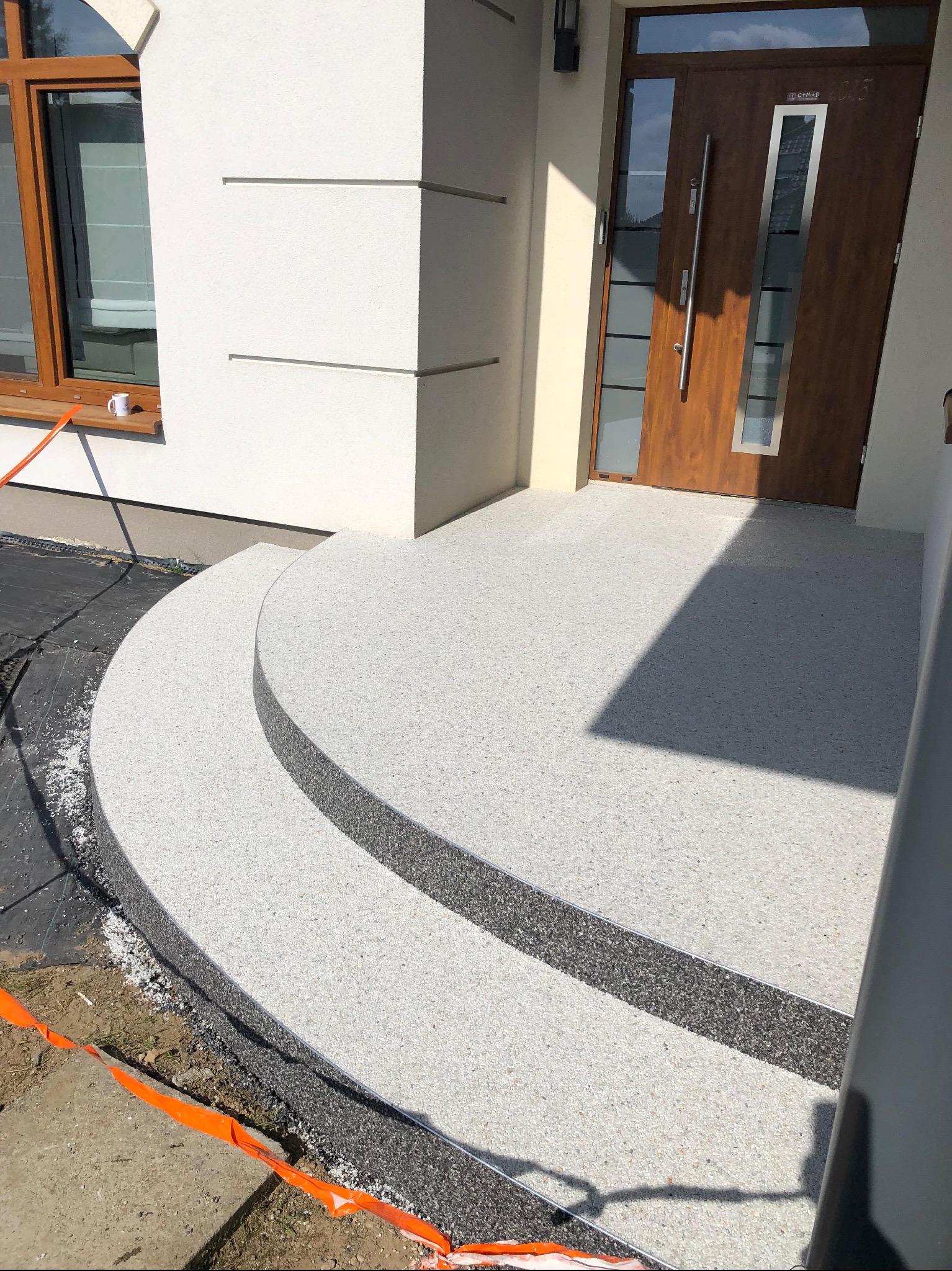 Wykończone schody wejściowe z jasnej, drobnoziarnistej posadzki żywicznej, z ciemnym obrzeżem. Widoczne drzwi wejściowe i fragment okna. Na zewnątrz taśma ostrzegawcza.