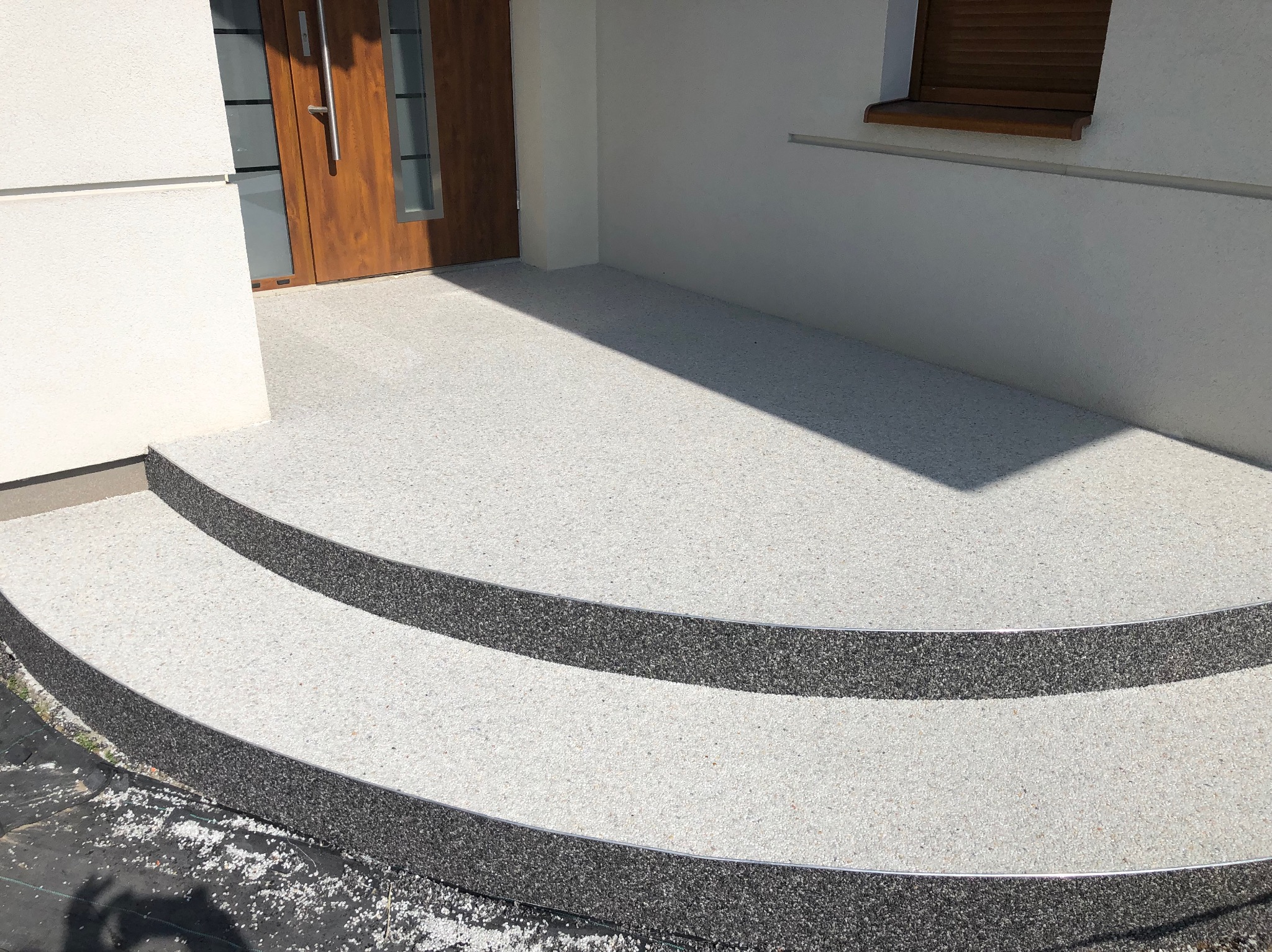 Jasna, drobnoziarnista posadzka żywiczna na schodach wejściowych do domu, z ciemnymi obrzeżami i metalowymi listwami wykończeniowymi. Widoczne drzwi wejściowe i fragment elewacji.