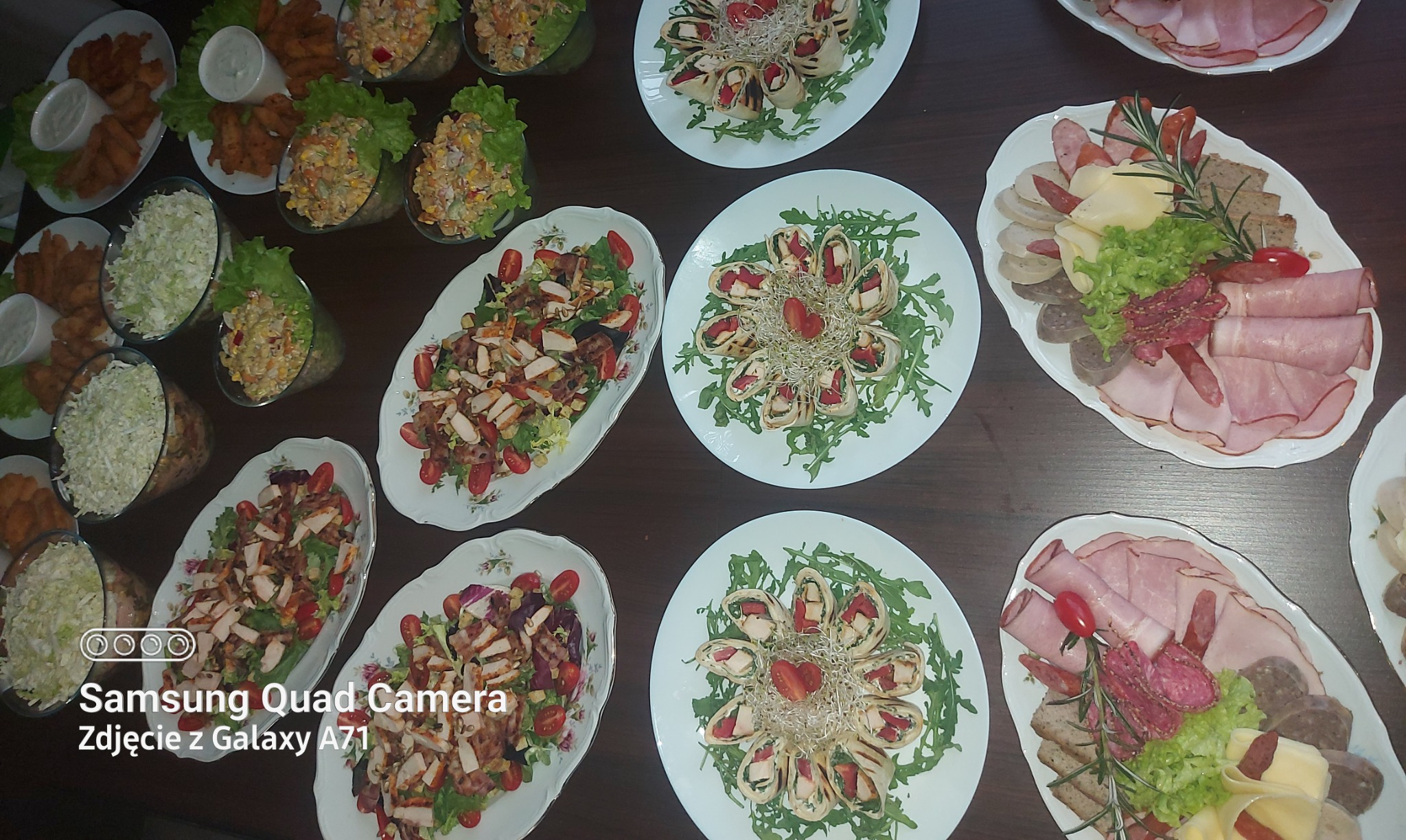 Stół z różnorodnymi potrawami cateringowymi: sałatki z kurczakiem i pomidorami na białych talerzach, tortille z warzywami i kiełkami, talerze wędlin i serów z rozmarynem, sałatki w szklankach...