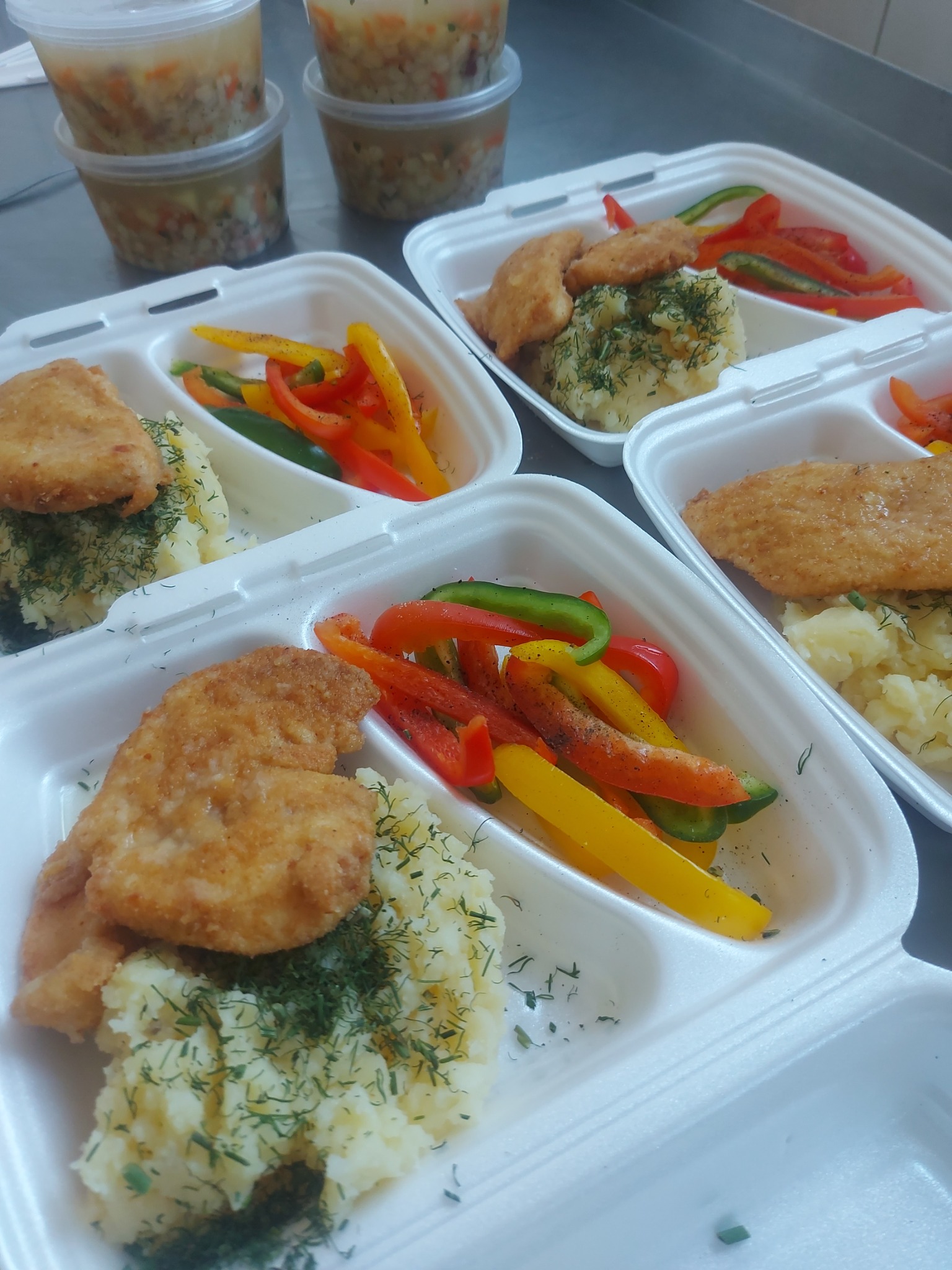 Gotowe zestawy obiadowe w białych tackach z podziałem na dania: kotlet, ziemniaki posypane koperkiem oraz papryka w trzech kolorach, przygotowane przez firmę z Kołbieli. W tle widoczne pojemniki...