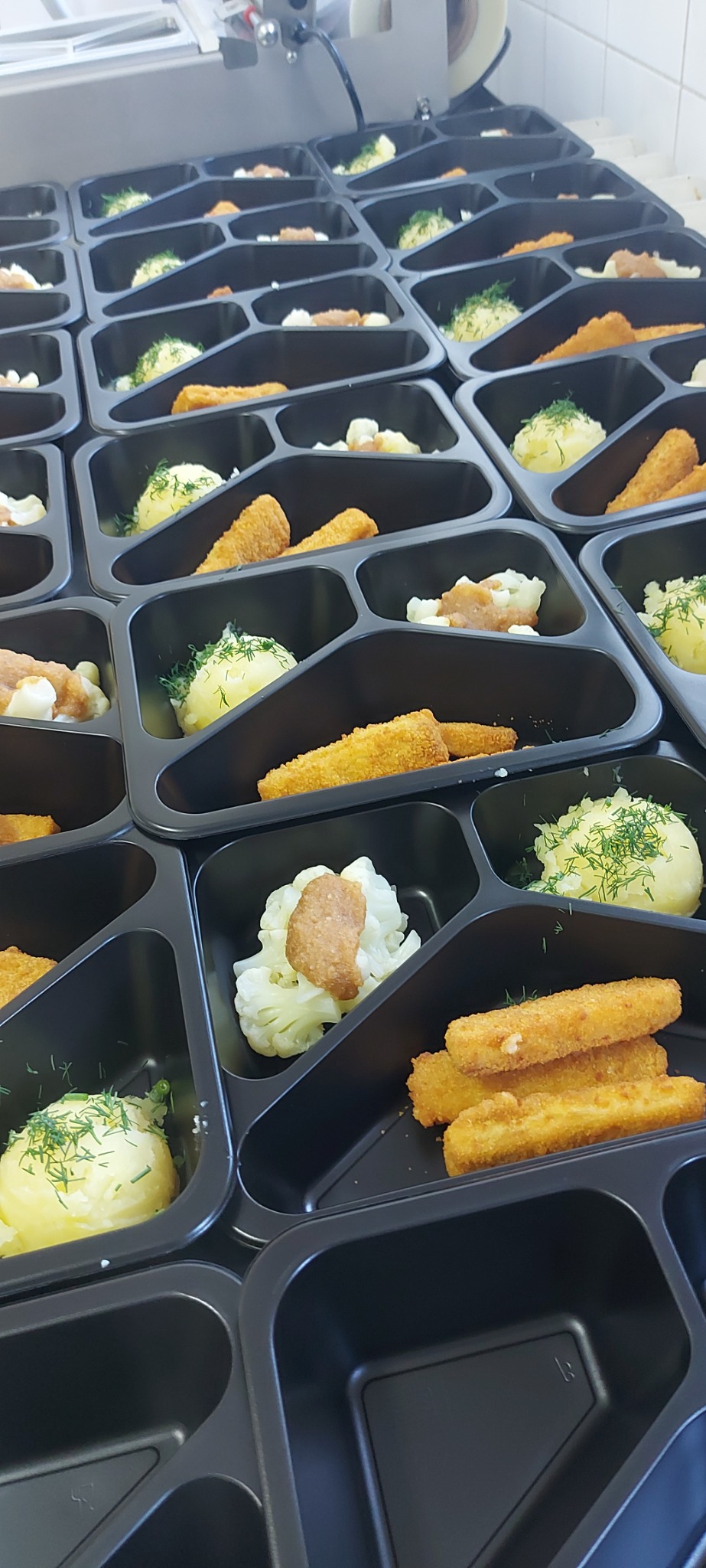 Przygotowane porcje obiadowe w czarnych, plastikowych pojemnikach: ryba w panierce, ziemniaki z koperkiem, kalafior z sosem, ułożone rzędami na białym blacie.