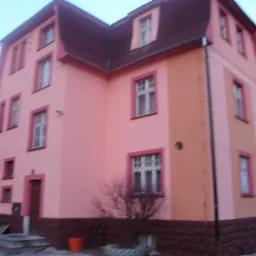 Dwukondygnacyjny budynek w Ludwikowicach KŁ. z docieploną i pomalowaną elewacją w odcieniach różu i pomarańczy, z ciemnoczerwoną podmurówką i dachem. Widoczne okna z białymi ramami.