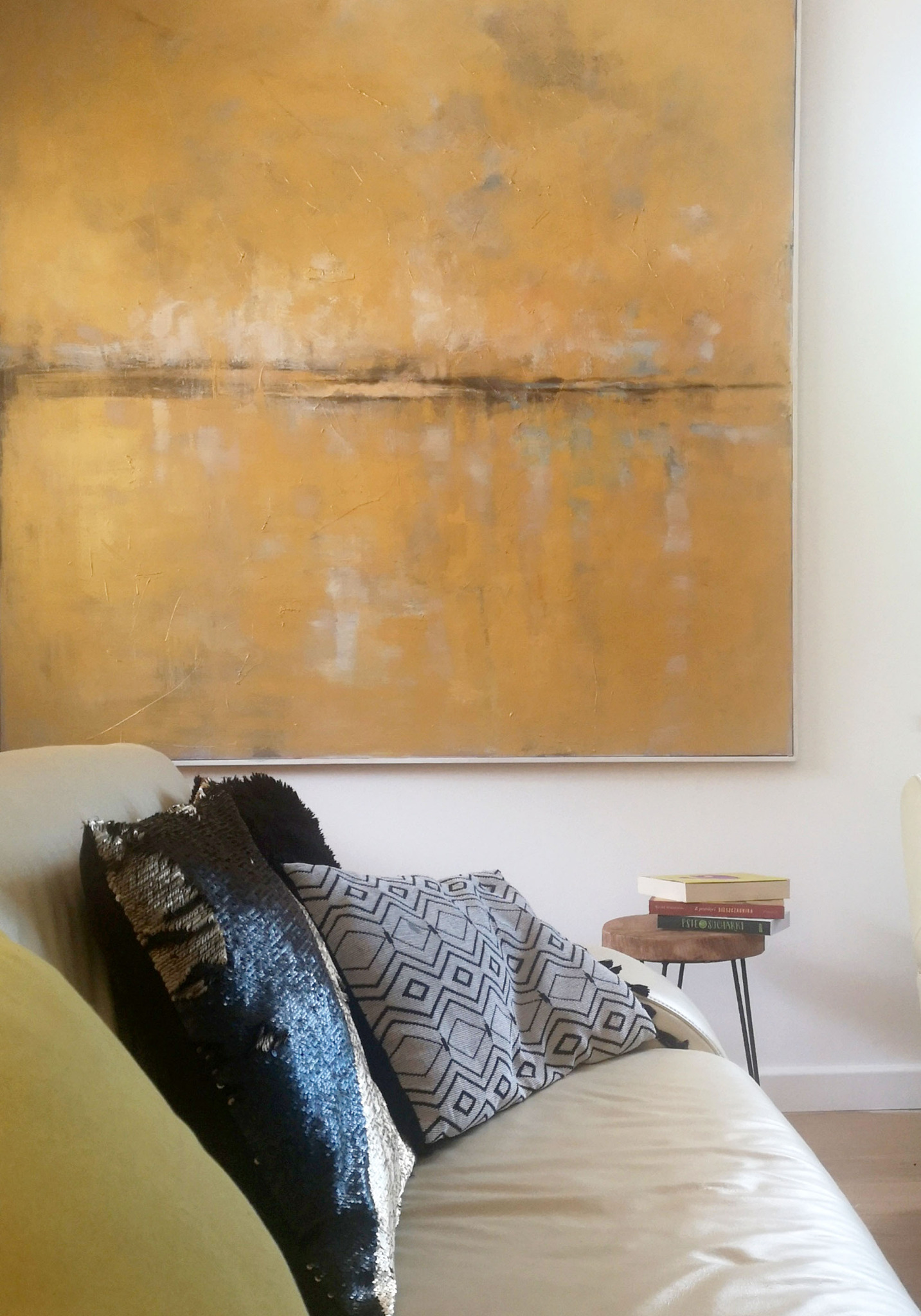 Abstrakcyjny obraz w odcieniach złota nad kremową sofą z dekoracyjnymi poduszkami w geometryczne wzory i cekiny, obok okrągły stolik z trzema książkami.