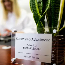 Tabliczka z napisem 'Kancelaria Adwokacka Adwokat Beata Kajewska tel.: 730 831 044' na biurku, obok doniczka z zieloną rośliną, w tle rozmazana postać kobiety.