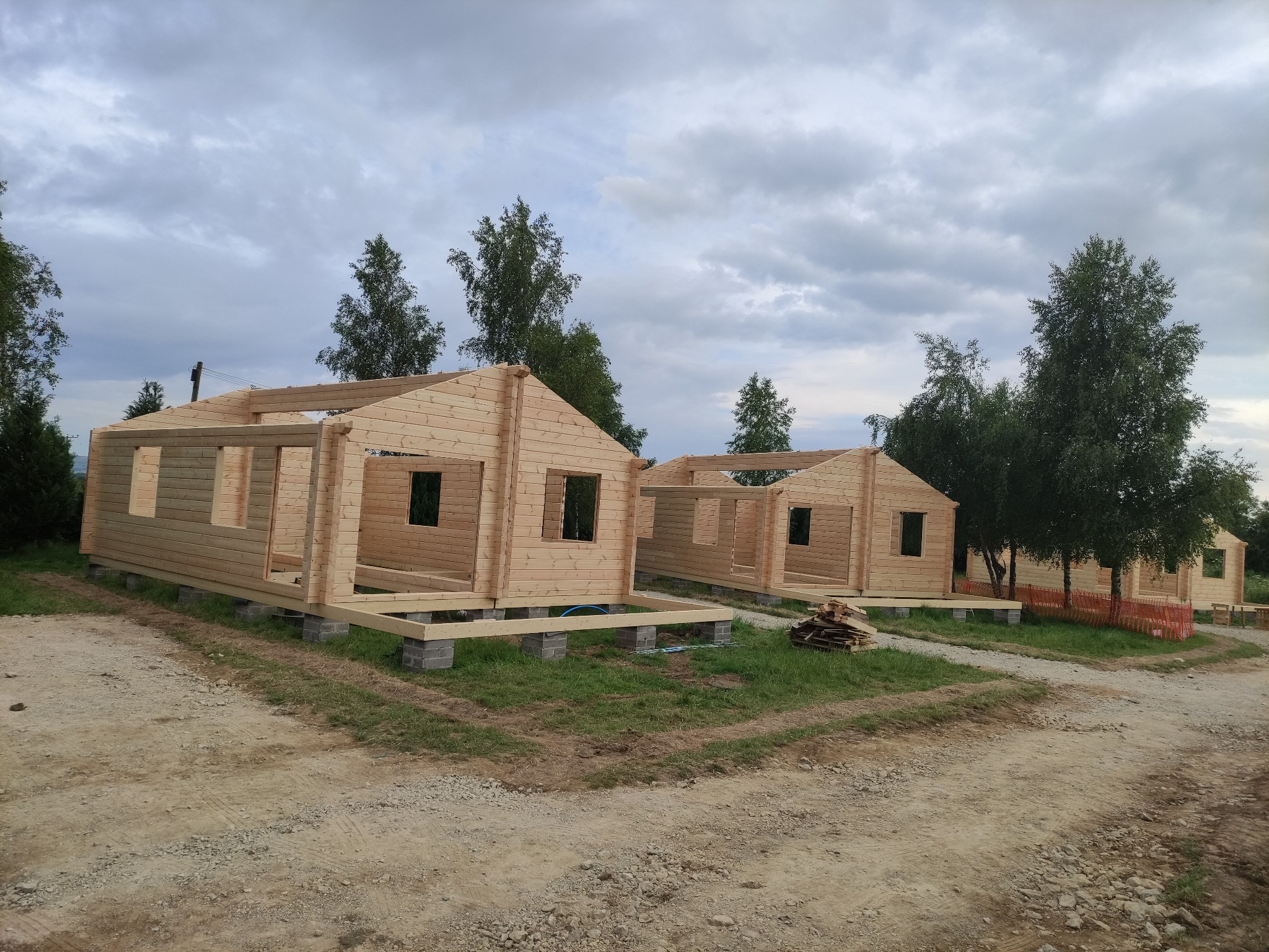 Wznoszenie szkieletowych domków letniskowych z jasnego drewna, widoczne fundamenty z betonowych bloczków i drewniana konstrukcja ścian z otworami okiennymi na tle pochmurnego nieba.