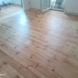 Świeżo wyszlifowana, jasna drewniana podłoga z widocznymi naturalnymi słojami i sękami, częściowo przygotowana do dalszej obróbki, z widocznym fragmentem okna i grzejnika w tle.