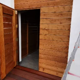 Zabudowa tarasowa z drewna z otwartymi drzwiczkami, widoczna drabina oparta o ścianę.