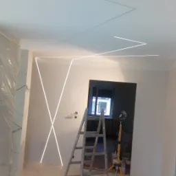 Wnętrze w trakcie montażu oświetlenia LED w kształcie geometrycznym na ścianie i suficie, drabina oparta o ścianę, podłoga zabezpieczona folią malarską.