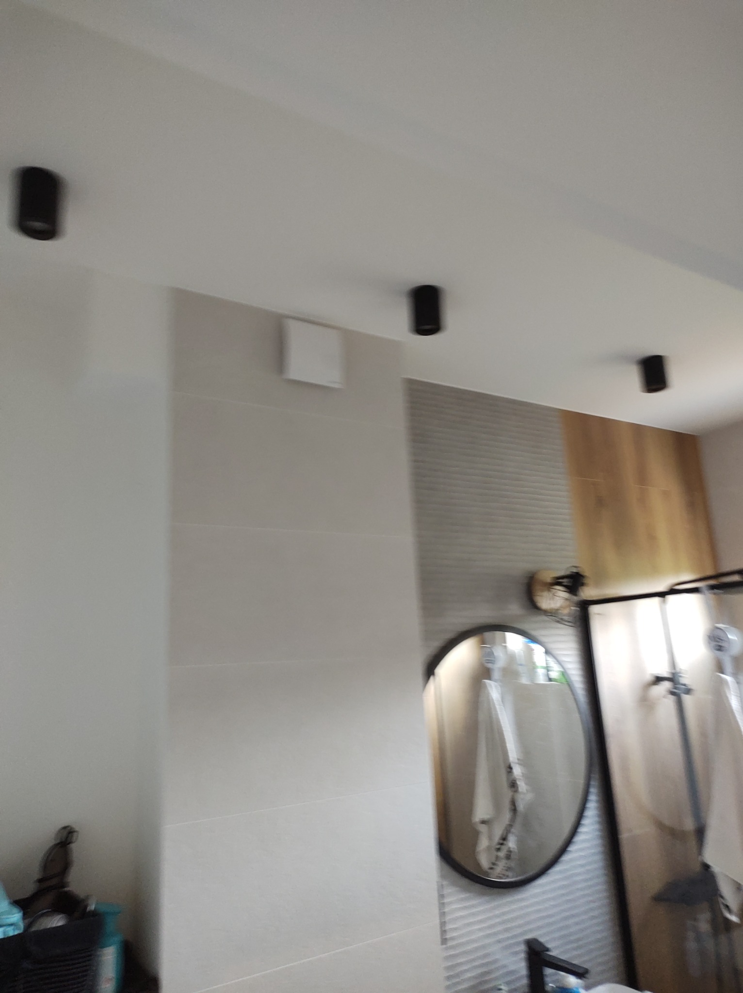 Wykończona łazienka z szarymi płytkami imitującymi drewno, okrągłym lustrem w czarnej ramie, czarnymi reflektorami sufitowymi oraz kabiną prysznicową z czarnymi elementami.