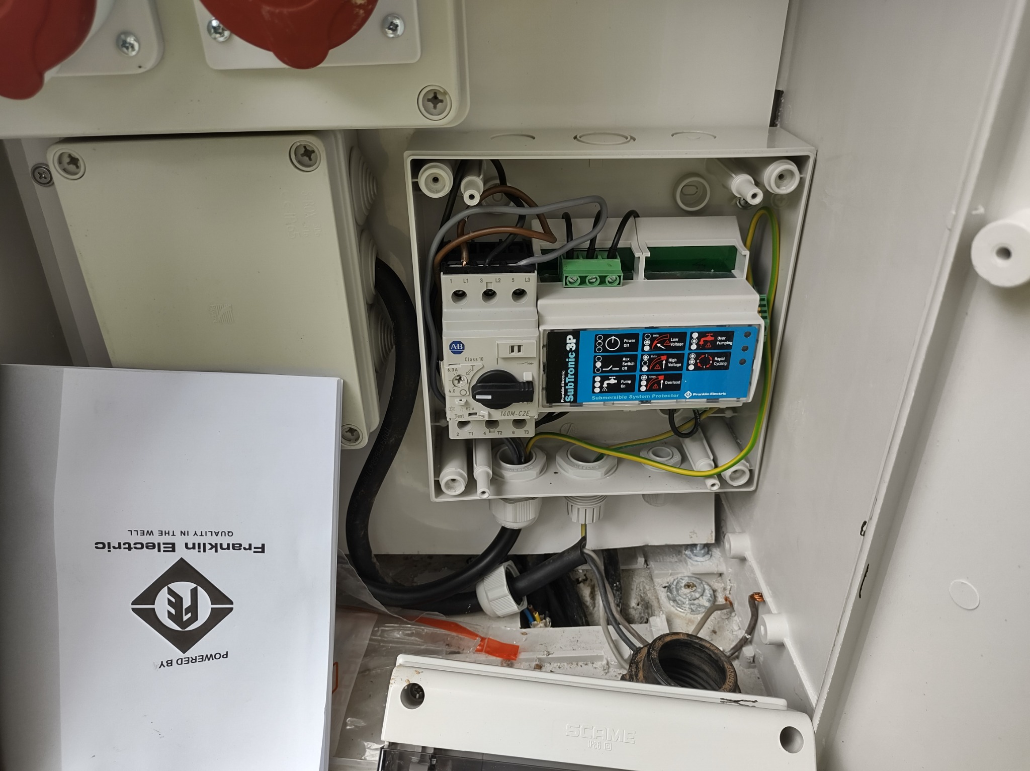 Otwarta skrzynka elektryczna z zamontowanym zabezpieczeniem pompy głębinowej SubTronic 3P firmy Franklin Electric, widoczne przewody, bezpiecznik, oraz instrukcja obsługi Franklin Electric.