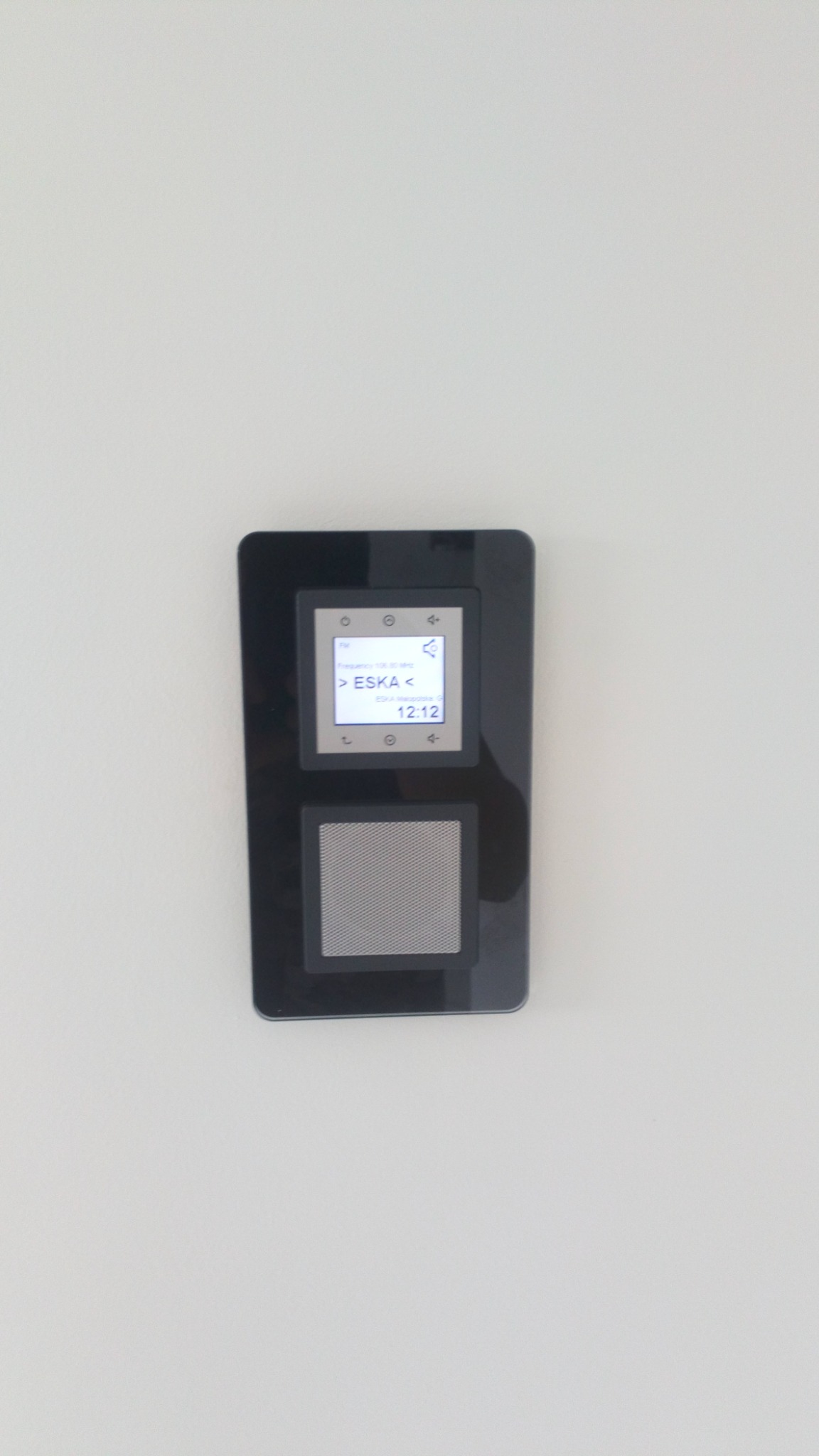 Ścienny panel sterowania inteligentnego domu, wyświetlający stację radiową ESKA, zamontowany na białej ścianie.