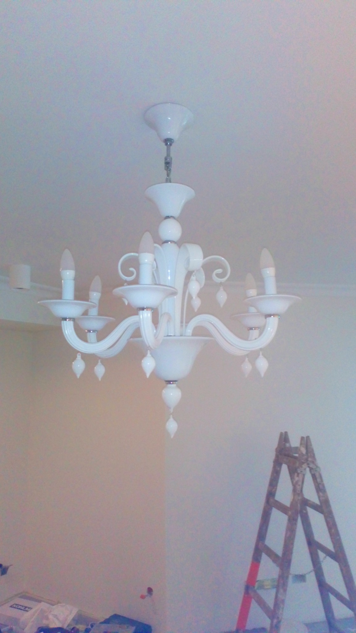 Biały, ozdobny żyrandol z żarówkami świecowymi zawieszony na suficie, z drewnianą drabiną opartą o ścianę w tle.