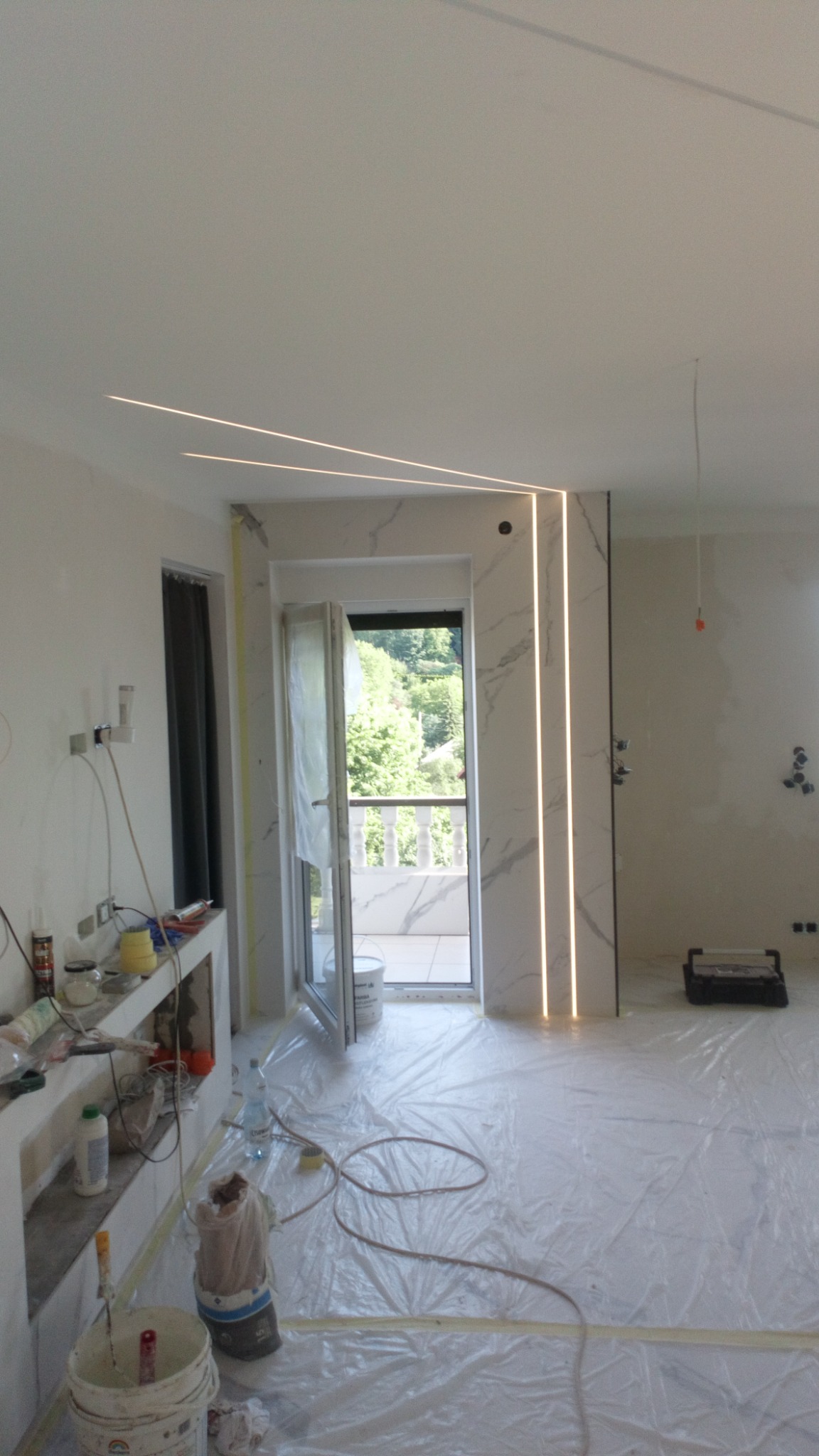 Wnętrze w trakcie remontu z widocznymi ledowymi liniami świetlnymi na suficie i ścianach, zabezpieczona folią podłoga, materiały budowlane i otwarte drzwi balkonowe z widokiem na zieleń.