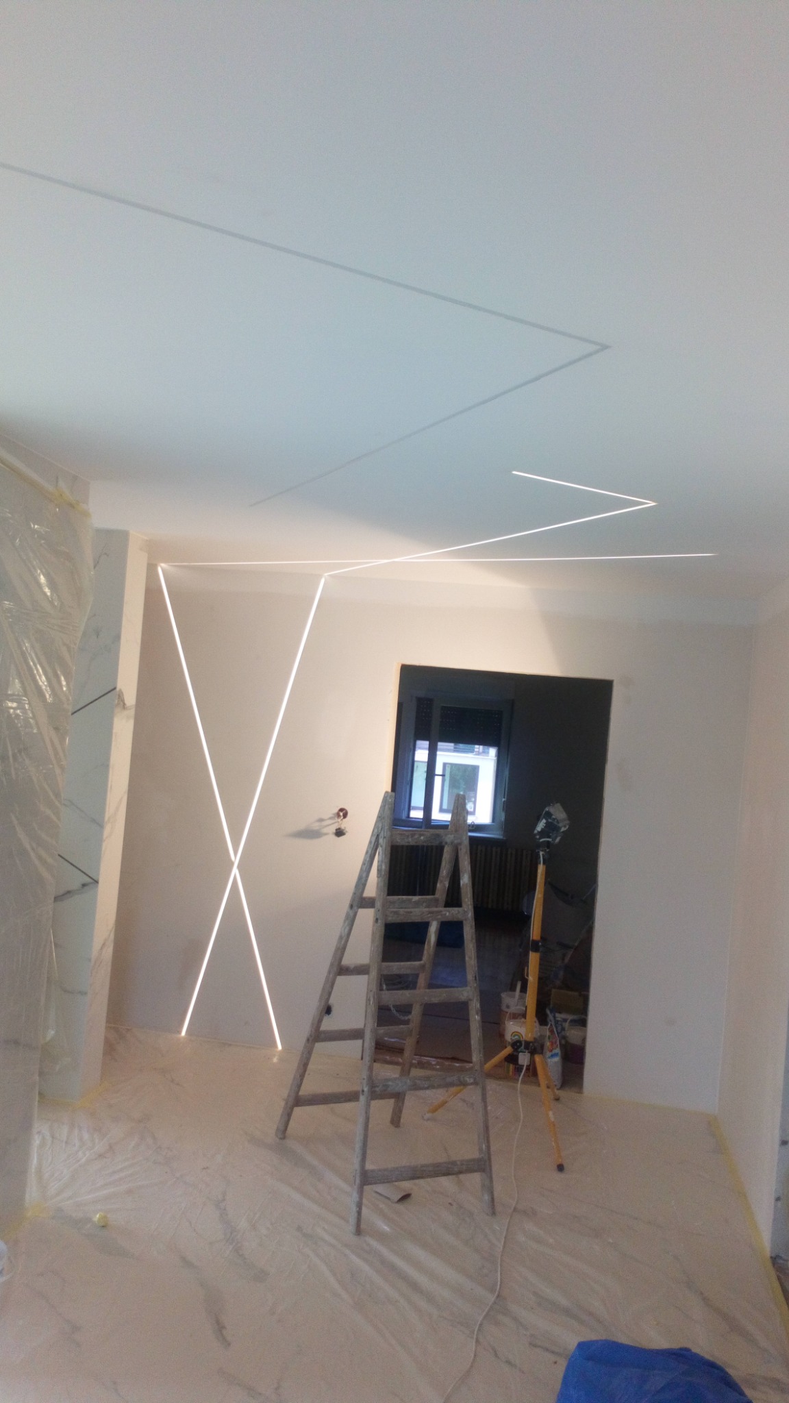 Wnętrze w trakcie montażu oświetlenia LED w kształcie geometrycznym na ścianie i suficie, drabina oparta o ścianę, podłoga zabezpieczona folią malarską.