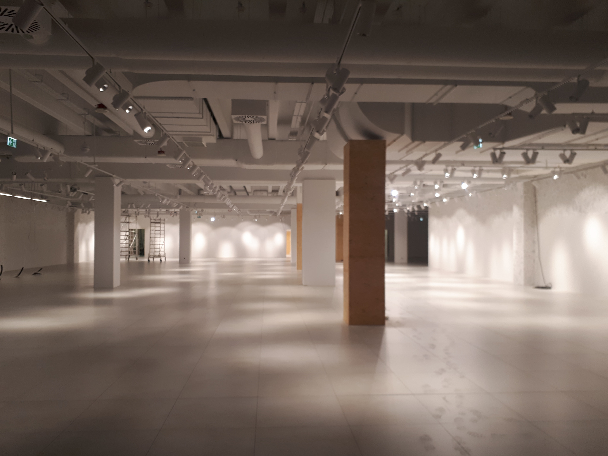 Przestronne, puste wnętrze biurowe z betonową podłogą, białymi ścianami i kolumnami, widoczne instalacje sufitowe oraz ślady na podłodze sugerujące prace wykończeniowe.