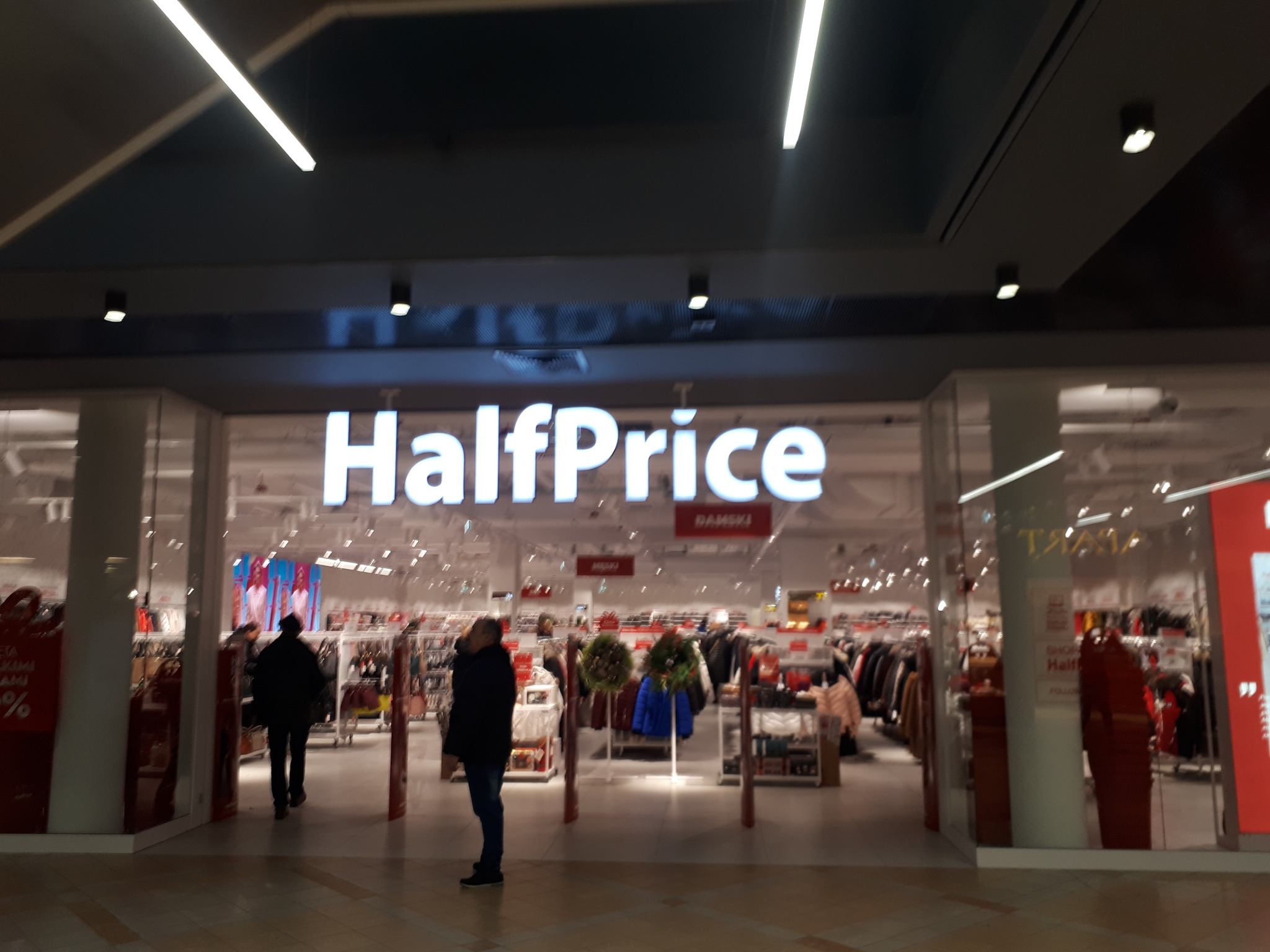 Wejście do sklepu HalfPrice z widocznymi osobami wchodzącymi i wychodzącymi, oświetlone neonowymi lampami sufitowymi, z ekspozycją odzieży i dekoracji świątecznych wewnątrz.
