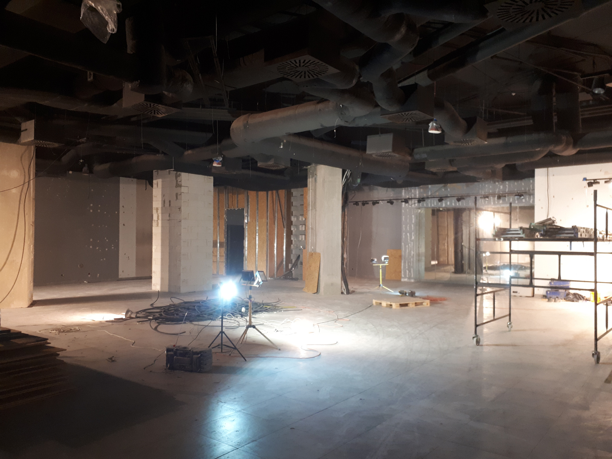 Przestronne wnętrze biurowe w trakcie prac wykończeniowych z widocznymi instalacjami pod sufitem, oświetleniem roboczym na statywie i przygotowanymi materiałami budowlanymi.