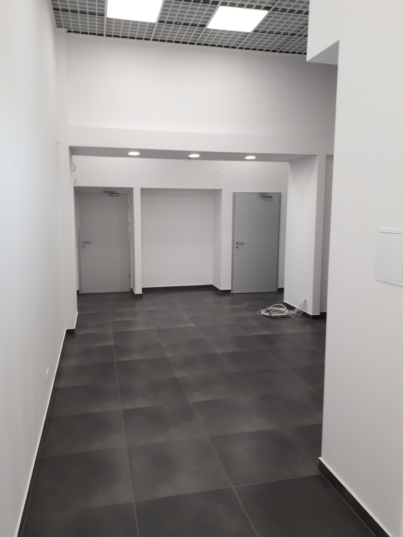 Korytarz biurowy z szarą podłogą z płytek, białymi ścianami, dwiema szarymi drzwiami i widocznymi kablami na podłodze.