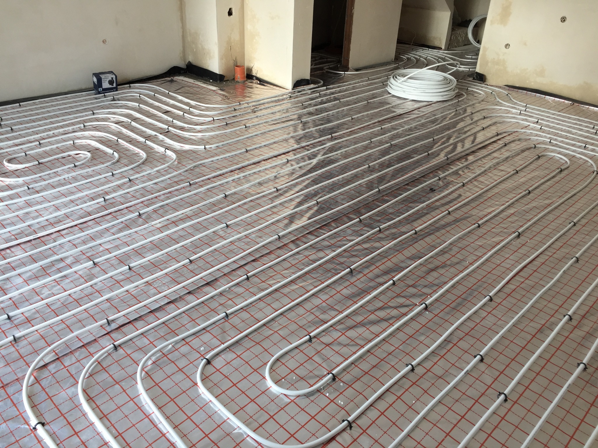 Rozłożona instalacja ogrzewania podłogowego na folii izolacyjnej, widoczny rozdzielacz i zwój rur.