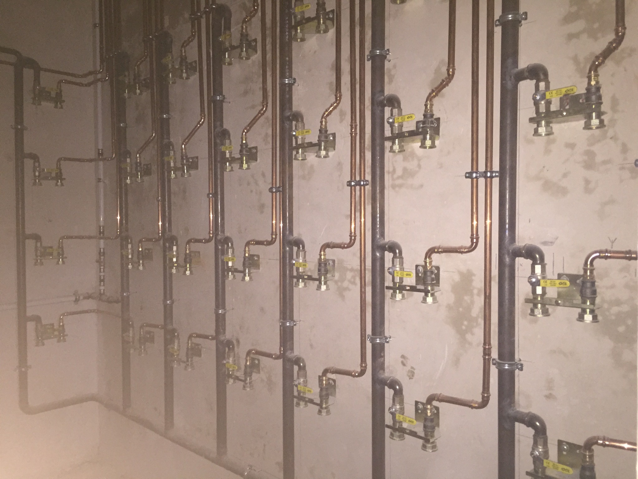 Rozbudowana instalacja wodna z zaworami, rurami miedzianymi i stalowymi, zamocowana do surowej, szarej ściany.