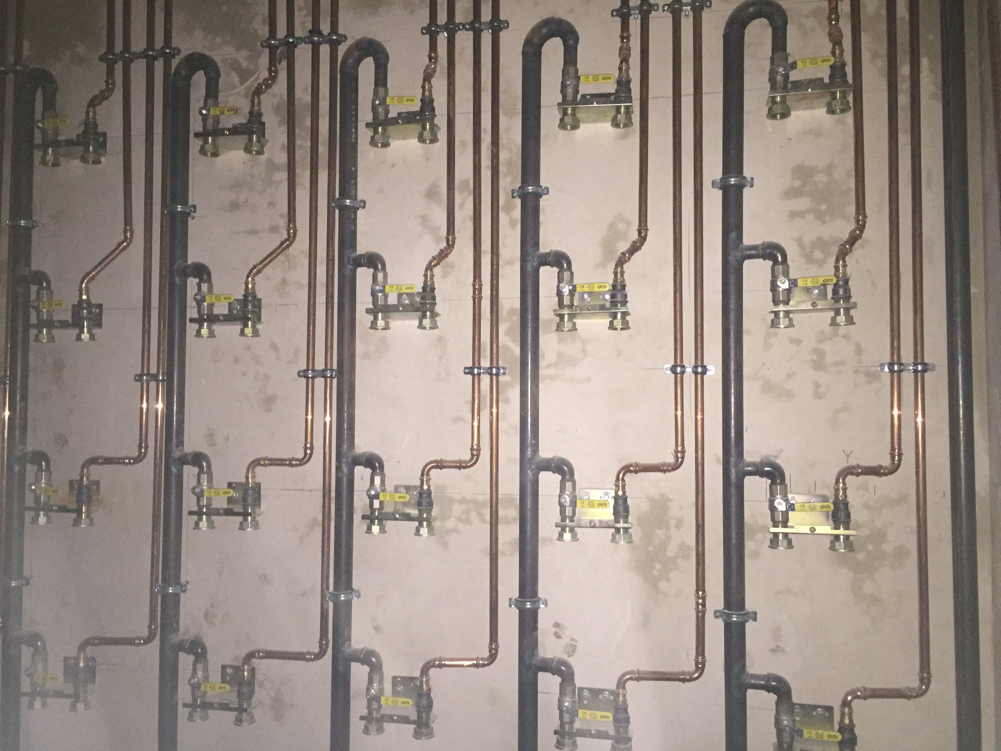 Rzędy pionowych rur miedzianych i czarnych, zamocowanych do ściany, z żółtymi zaworami i elementami mocującymi.