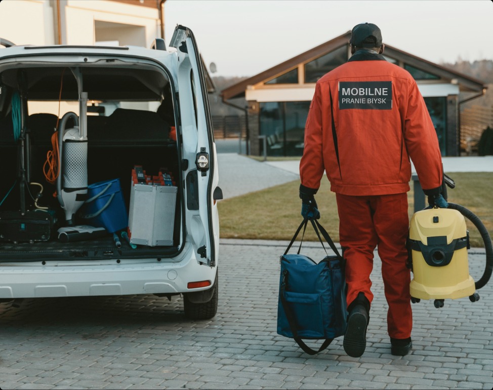 Pracownik w czerwonym kombinezonie z napisem 'Mobilne Pranie Błysk' idzie po brukowanej ścieżce w kierunku otwartego bagażnika białego samochodu dostawczego, niosąc torbę i żółty odkurzacz, z tyłu...