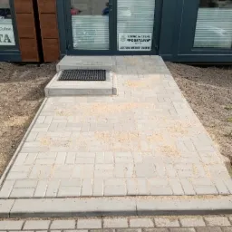 Nowo ułożony chodnik z jasnoszarej kostki brukowej prowadzący do wejścia agencji celnej, z widocznymi pozostałościami piasku na powierzchni