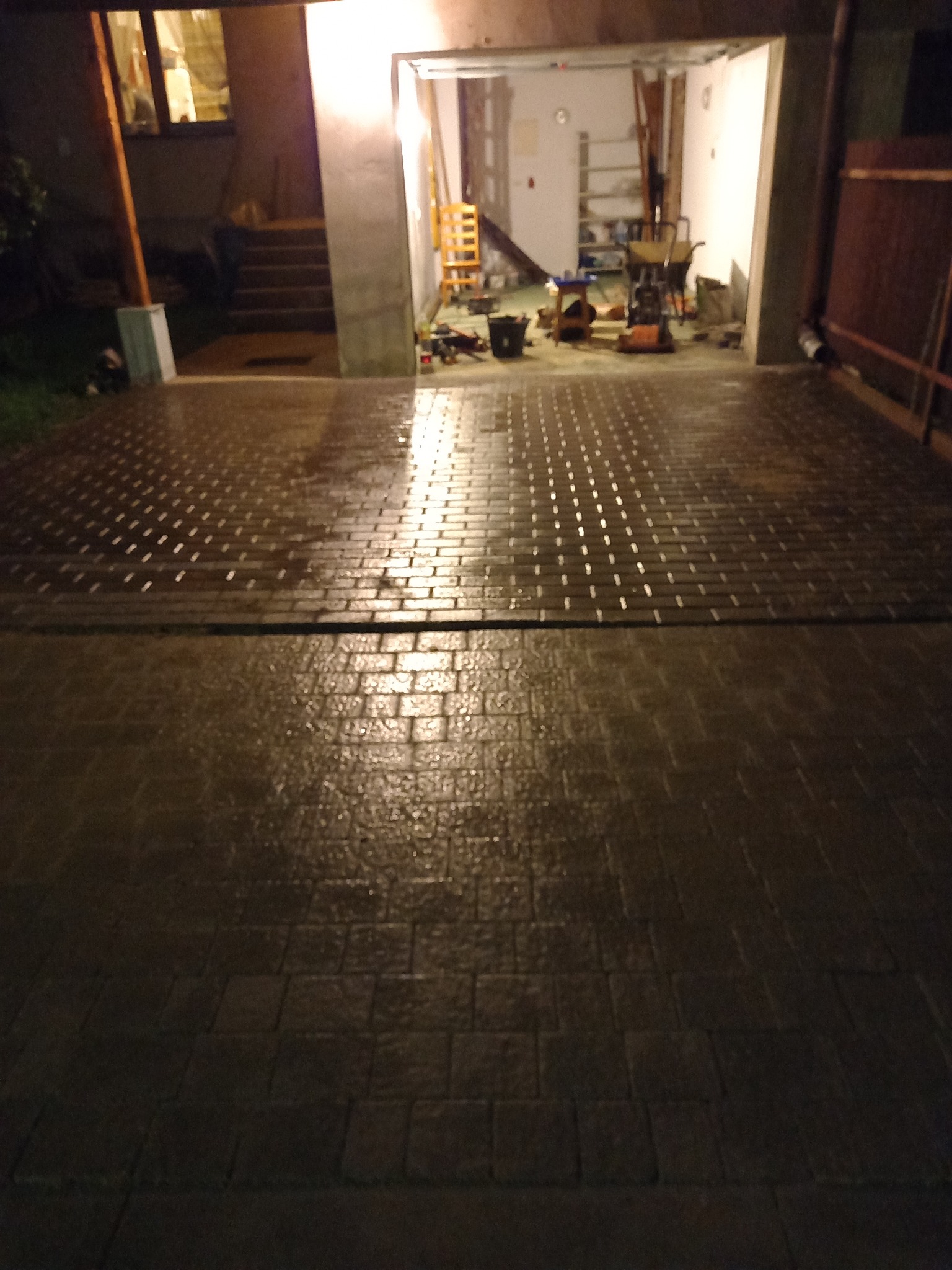 Nowo ułożona, mokra kostka brukowa w Łodzi, odbijająca światło z otwartego garażu, w tle widoczne narzędzia i sprzęt budowlany.