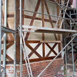 Alpex sp. z o.o. - Fragment ściany szachulcowej w trakcie renowacji, widoczne rusztowanie, odnowione fragmenty z jasną zaprawą i ciemnym drewnem oraz fragmenty ceglanej podmurówki z ubytkami tynku.