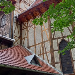 Alpex sp. z o.o. - Fragment zabytkowej kamienicy z muru pruskiego, widok z dołu na elewację z drewnianymi belkami i wypełnieniami o fakturze tynku, czerwony dach z lukarną i zielone liście drzew na pierwszym planie.