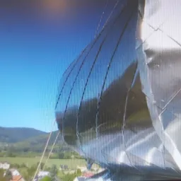 Nietypowa, błyszcząca elewacja budynku z metalowych paneli o zaokrąglonym kształcie, odbijająca błękitne niebo, na tle zielonych wzgórz i zabudowań.