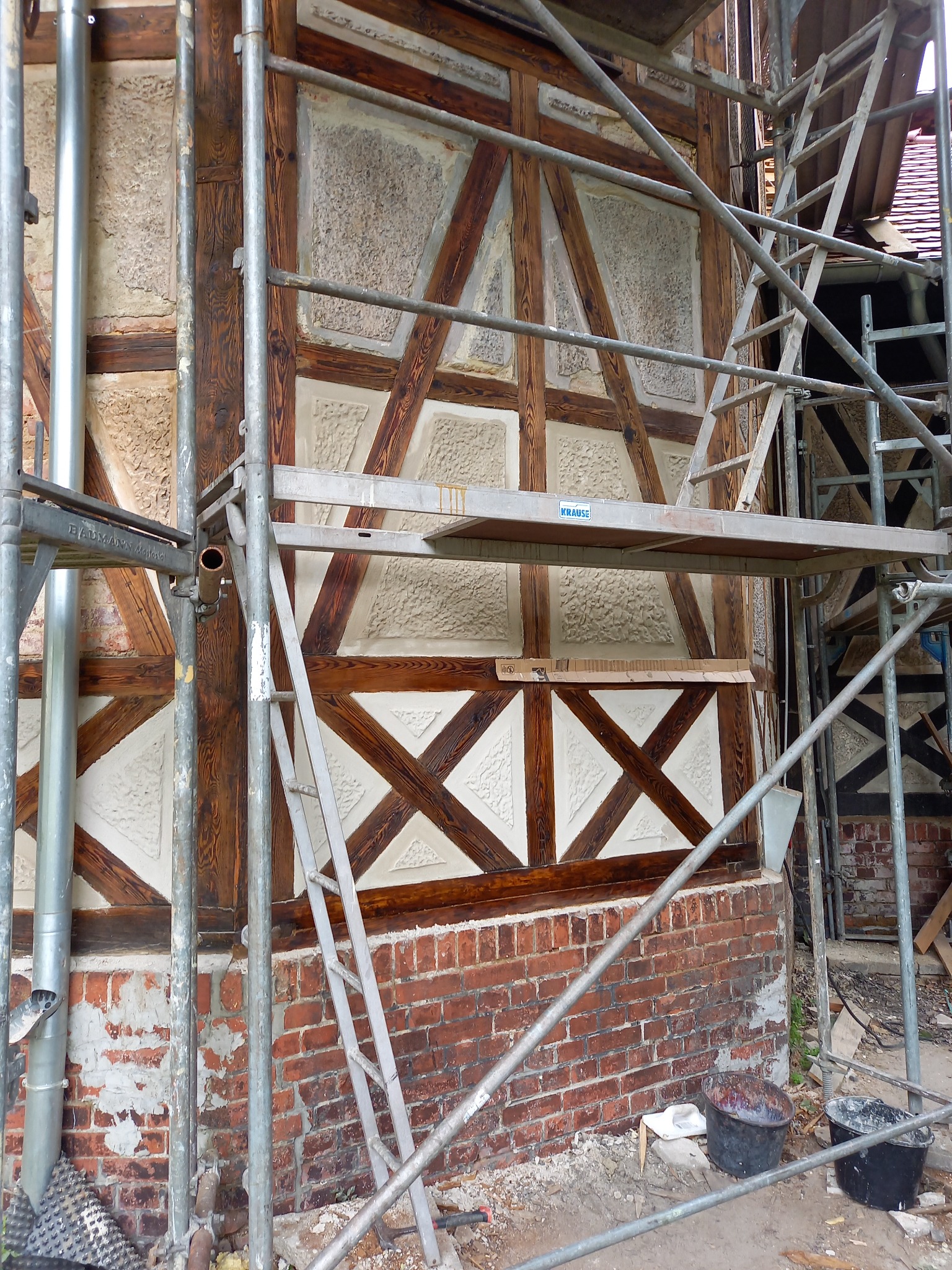 Fragment ściany szachulcowej w trakcie renowacji, widoczne rusztowanie, odnowione fragmenty z jasną zaprawą i ciemnym drewnem oraz fragmenty ceglanej podmurówki z ubytkami tynku.