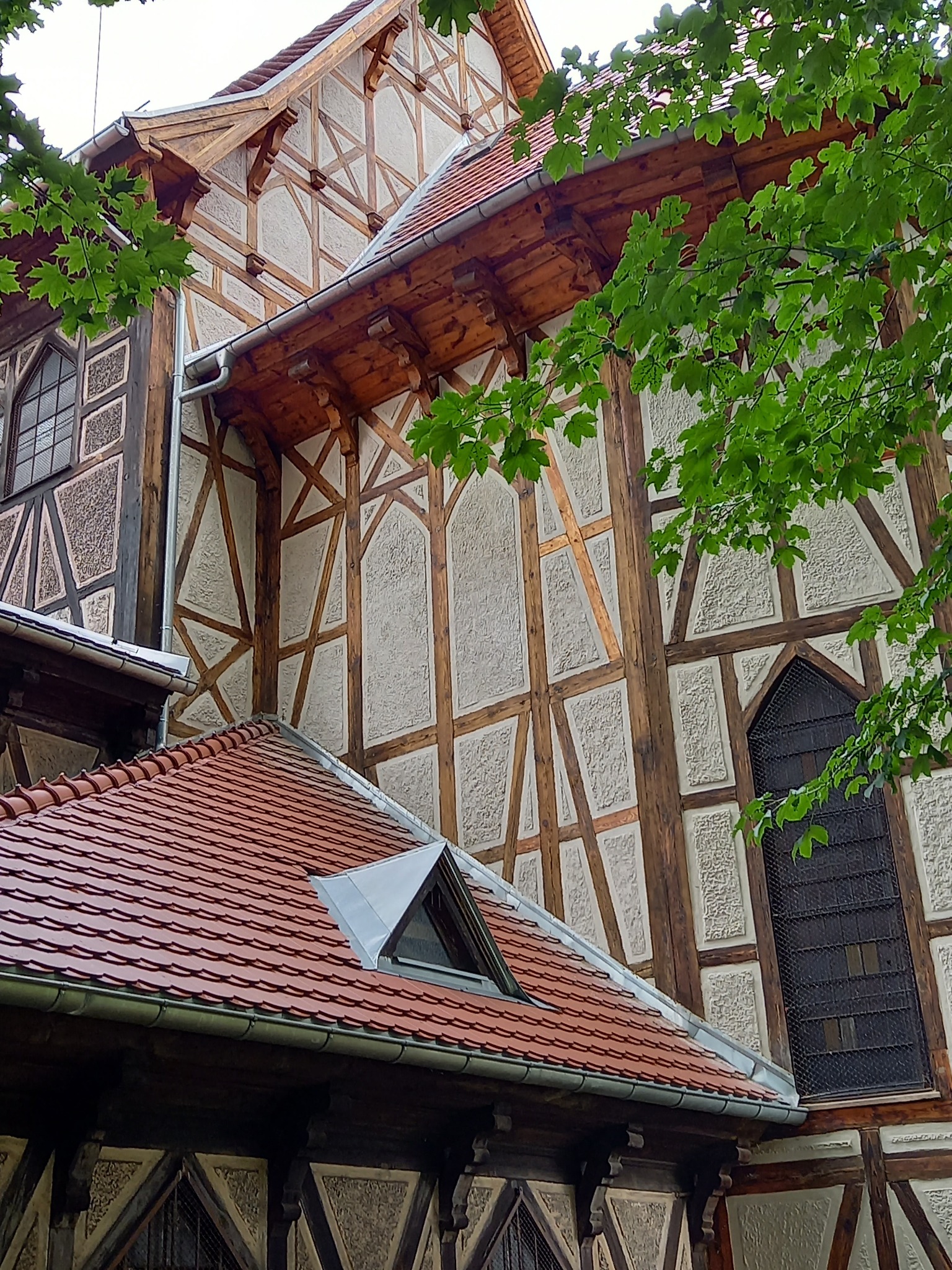 Fragment zabytkowej kamienicy z muru pruskiego, widok z dołu na elewację z drewnianymi belkami i wypełnieniami o fakturze tynku, czerwony dach z lukarną i zielone liście drzew na pierwszym planie.