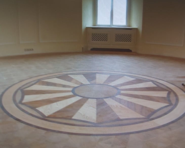 Wnętrze z parkietem w kształcie rozety, wykonanym z różnych gatunków drewna, z widocznym grzejnikiem pod oknem.