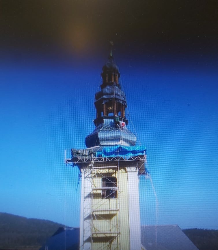 Wieża kościoła w trakcie renowacji z użyciem rusztowań i plandek ochronnych, widoczni pracownicy na górnych poziomach.