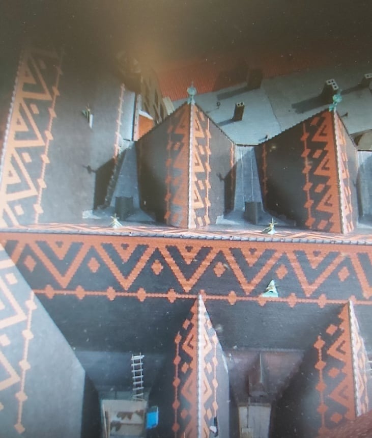 Widok z góry na odrestaurowaną kamienicę z charakterystycznym wzorem geometrycznym w kolorze terakoty na elewacji i dachu; widoczne elementy dekoracyjne na szczytach budynków.