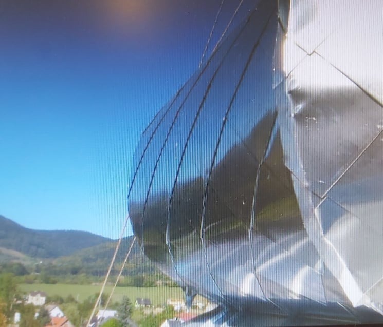 Nietypowa, błyszcząca elewacja budynku z metalowych paneli o zaokrąglonym kształcie, odbijająca błękitne niebo, na tle zielonych wzgórz i zabudowań.