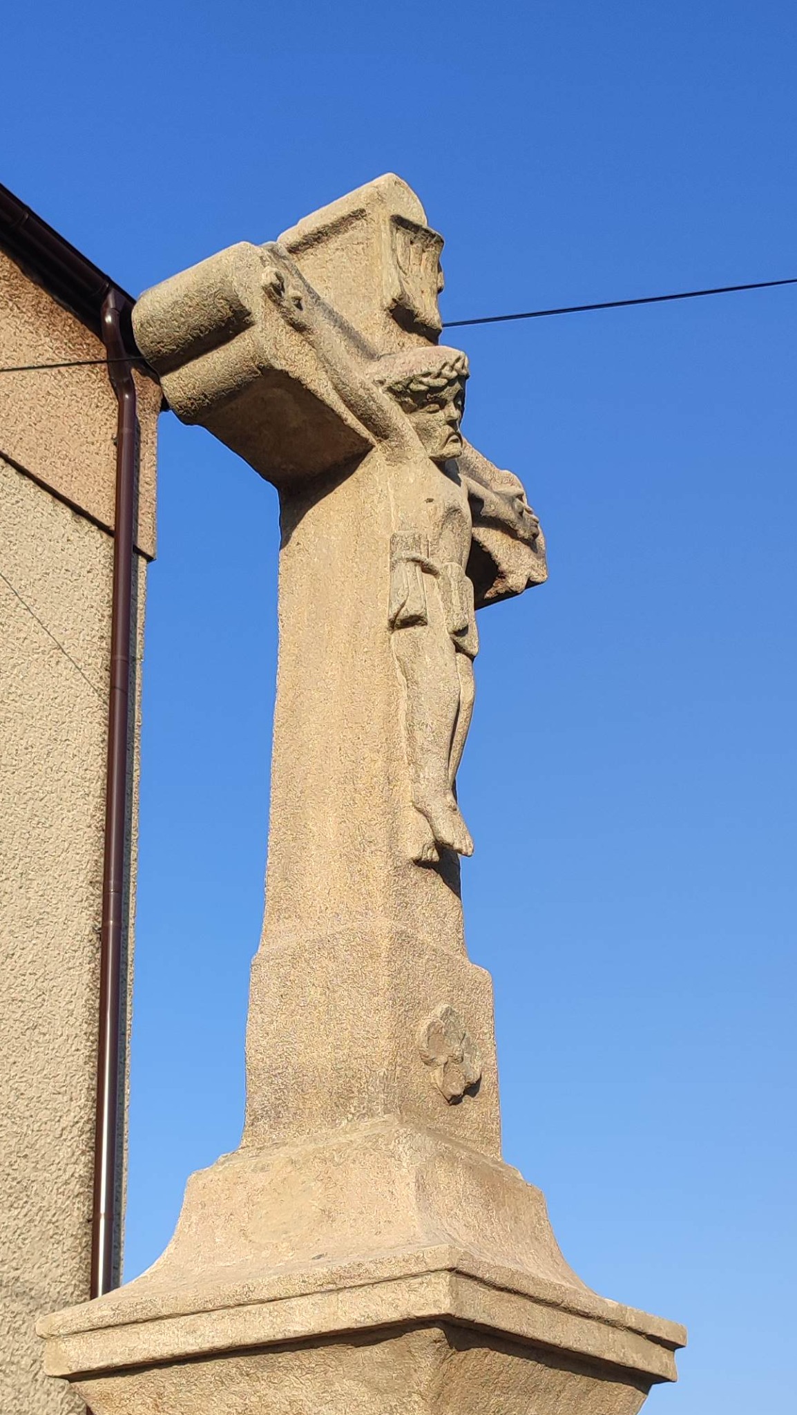 Kamienna kapliczka z figurą Chrystusa na tle błękitnego nieba, widoczna z dołu. Detal architektoniczny z elementami zdobniczymi i fragmentem budynku obok.