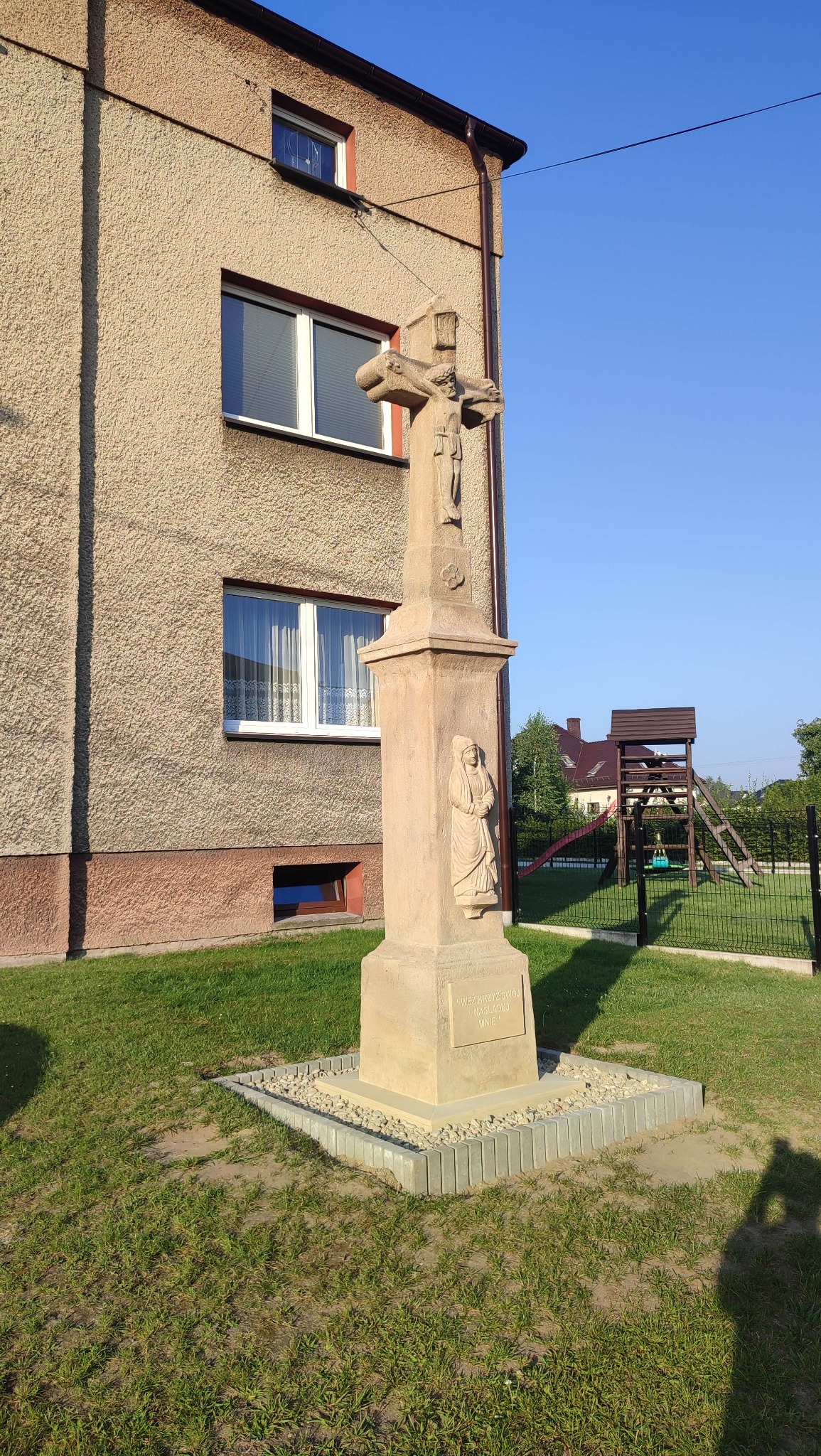 Kamienny krzyż z płaskorzeźbami na tle budynku z elewacją typu baranek i placu zabaw. Jasne, słoneczne światło.