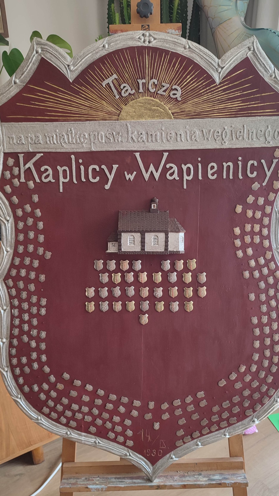 Pamiątkowa tarcza z modelem Kaplicy w Wapienicy i herbami, rok 1930, na drewnianym stojaku. Czerwone tło, srebrne obramowanie i napisy. Detal.