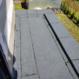 Widok z góry na płaski dach pokryty papą termozgrzewalną, widoczny fragment ściany budynku, w tle szopa i stos drewna na działce z zielenią.