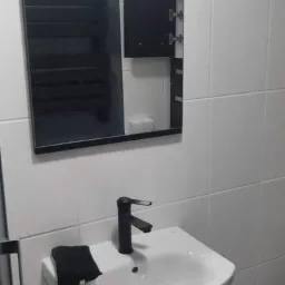 Nowoczesna umywalka z czarną szafką i baterią, nad nią lustro z widocznym grzejnikiem łazienkowym w odbiciu, białe płytki na ścianie.