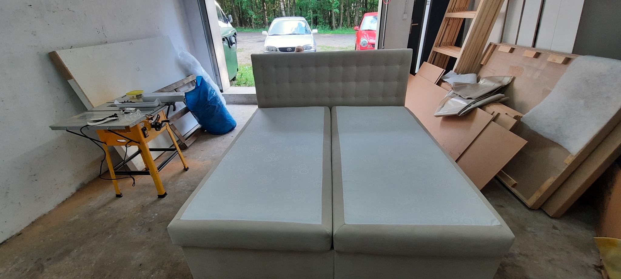 Dwa moduły łóżka tapicerowanego z pikowanym zagłówkiem w warsztacie, widoczny stół z piłą, materiały i samochody w tle.