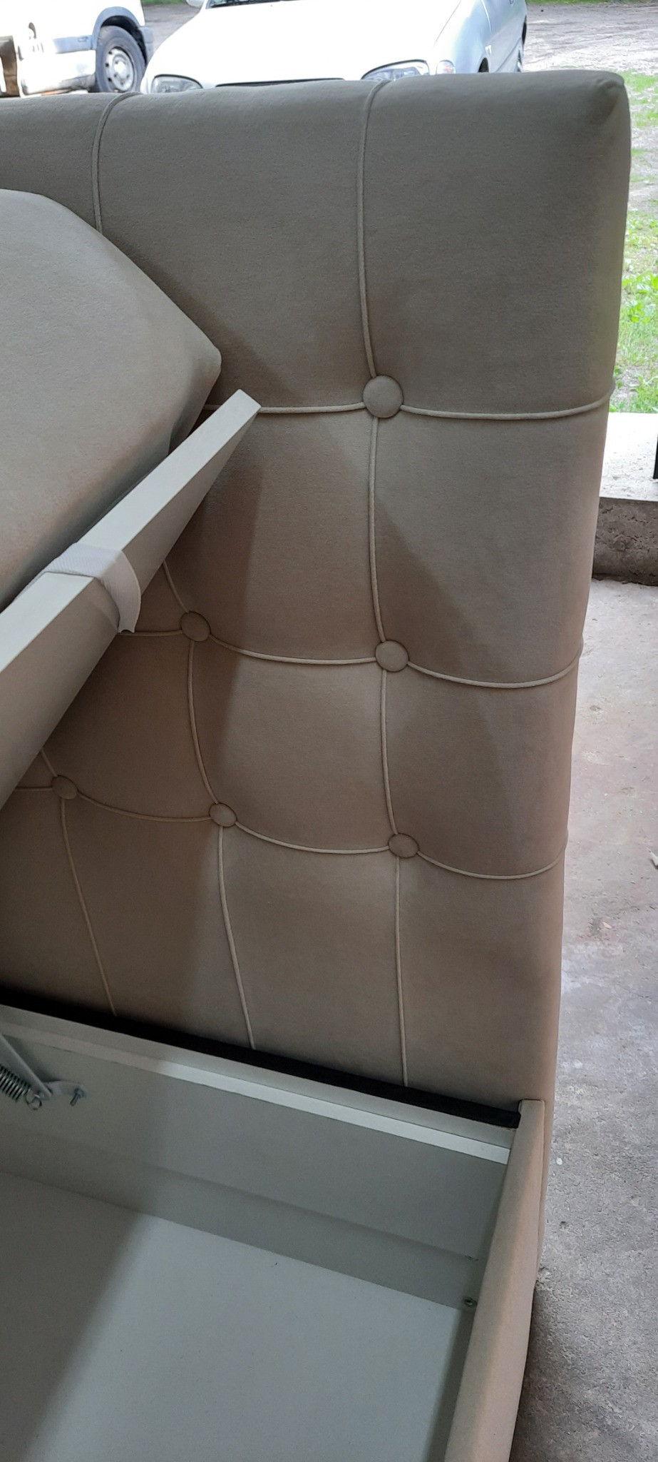 Beżowe tapicerowane wezgłowie łóżka z pikowaniem i pojemnikiem na pościel w trakcie otwierania, widoczne sprężyny mechanizmu podnoszenia.