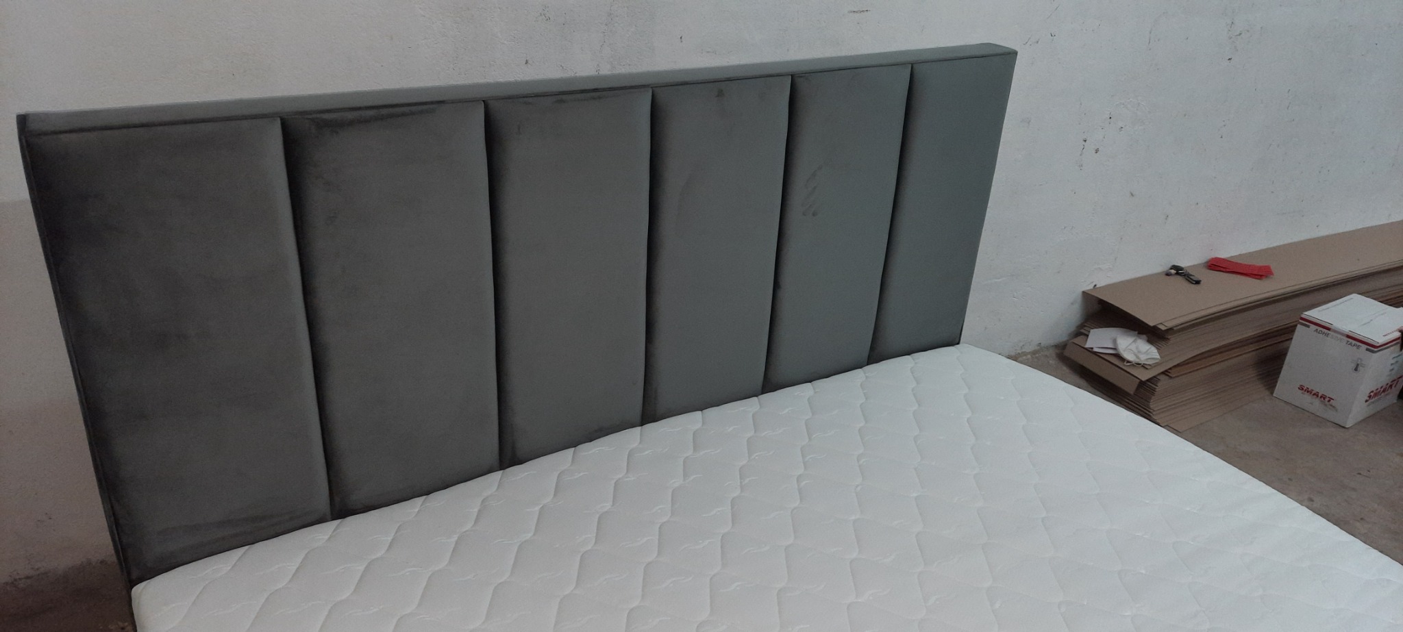 Szare, tapicerowane wezgłowie łóżka z pionowymi panelami, biały materac, w tle sterta kartonów i narzędzia.