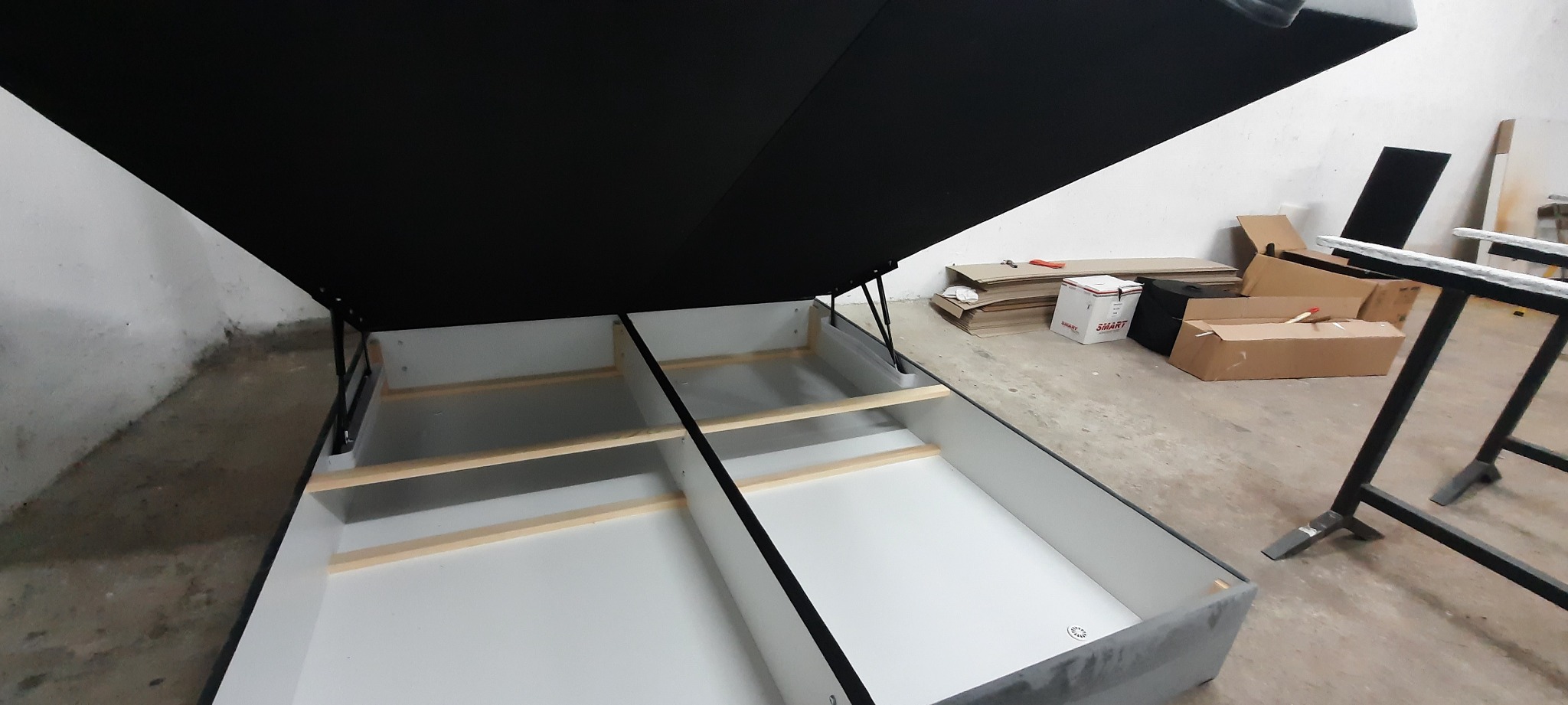Otwarta rama łóżka tapicerowanego z podnośnikiem, widoczne wewnętrzne przegrody i podłoga skrzyni, w tle kartony i narzędzia w warsztacie.