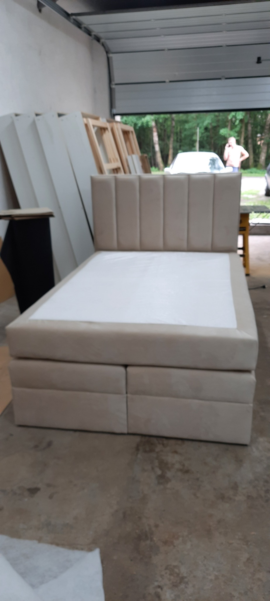 Beżowe łóżko tapicerowane z wezgłowiem w pionowe pasy, sfotografowane w warsztacie na tle otwartej bramy garażowej i elementów drewnianych konstrukcji.