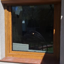 Tomasz Chałat SET - Brązowa rama okienna z zamontowaną roletą zewnętrzną, widoczna z zewnątrz budynku, z terakotowym parapetem i odbiciem w szybie.